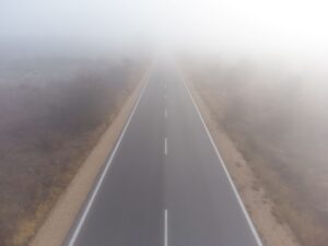 A straight paved road extends into dense fog that obscures the horizon. White lane markings are visible on the asphalt, with gravel shoulders and sparse dry grass along both sides.