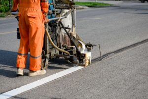 A worker in an orange reflective uniform operates a road marking machine that applies a fresh white line of paint onto an asphalt roadway. The machine releases a steady stripe as the worker guides it forward.