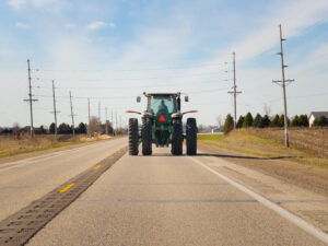 Farm tractor with slow-moving vehicle (SMV) signage traveling on a rural two-lane road through farmland.